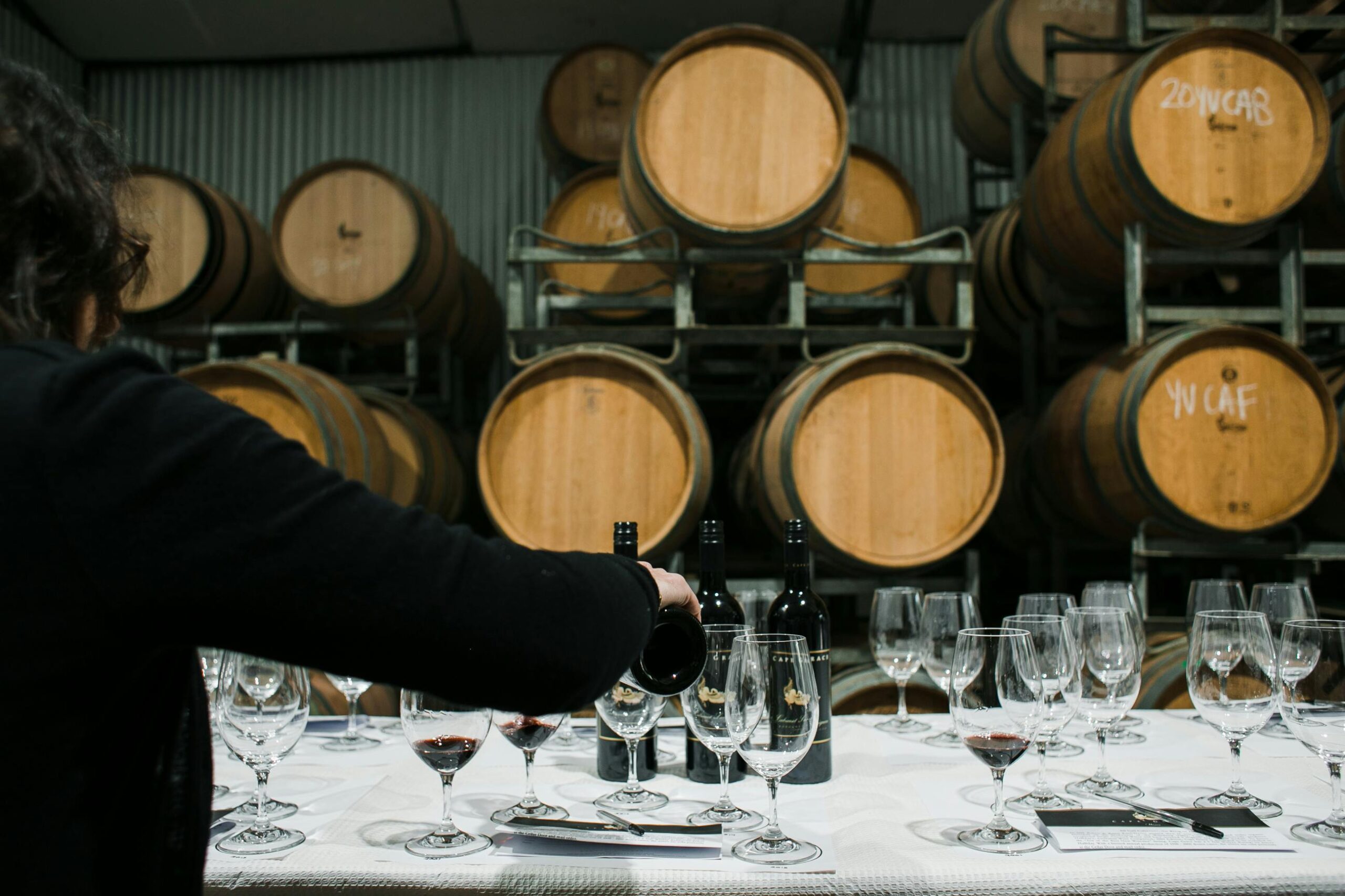 Woman pouring wine to glass while surrounded by wine barrels in a wine cellar.